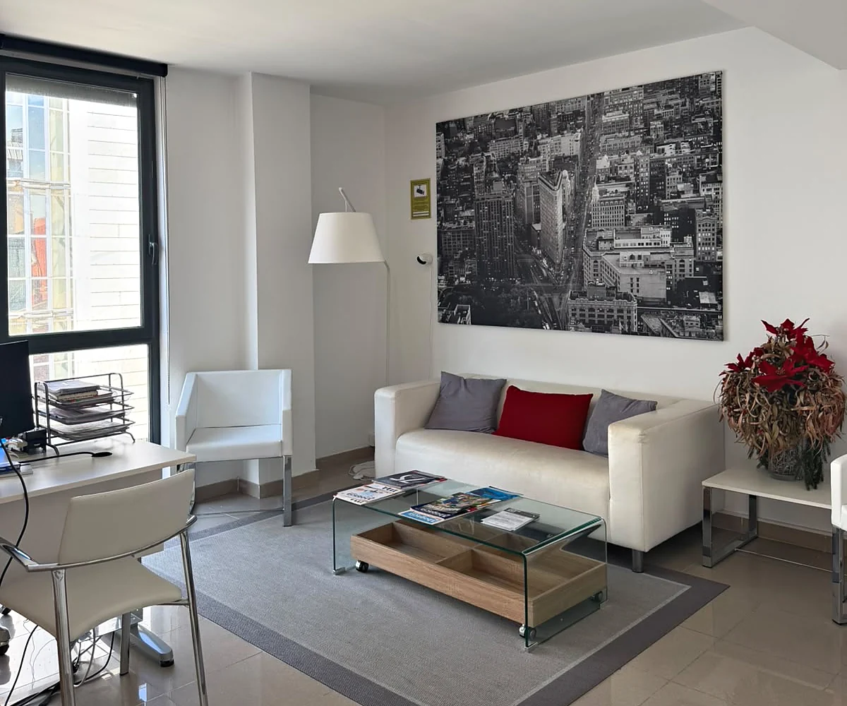 Zona de espera del despacho con sofá blanco, sillón y mesa baja de cristal.
